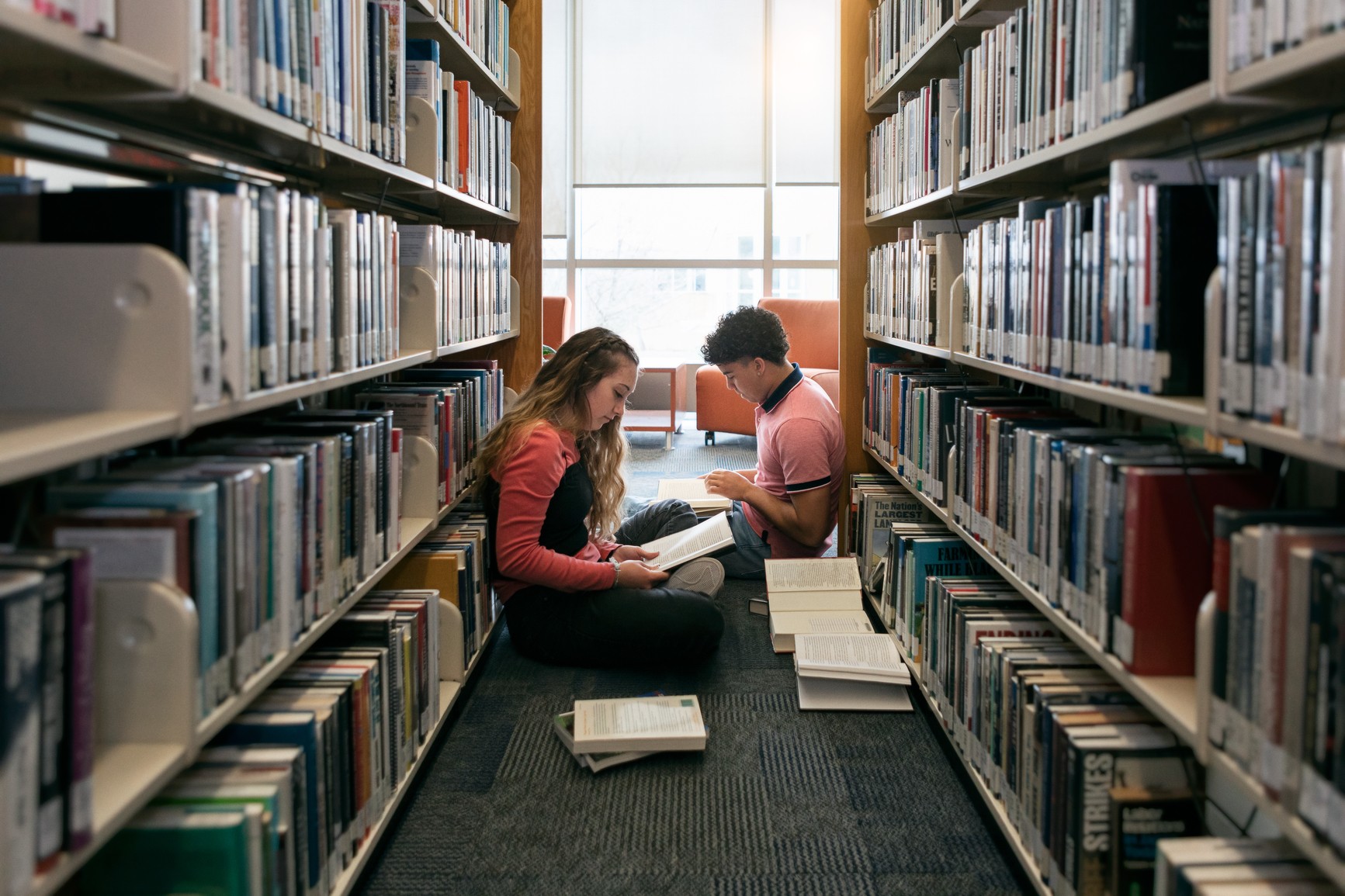 Two students reading on the floor between library shelves